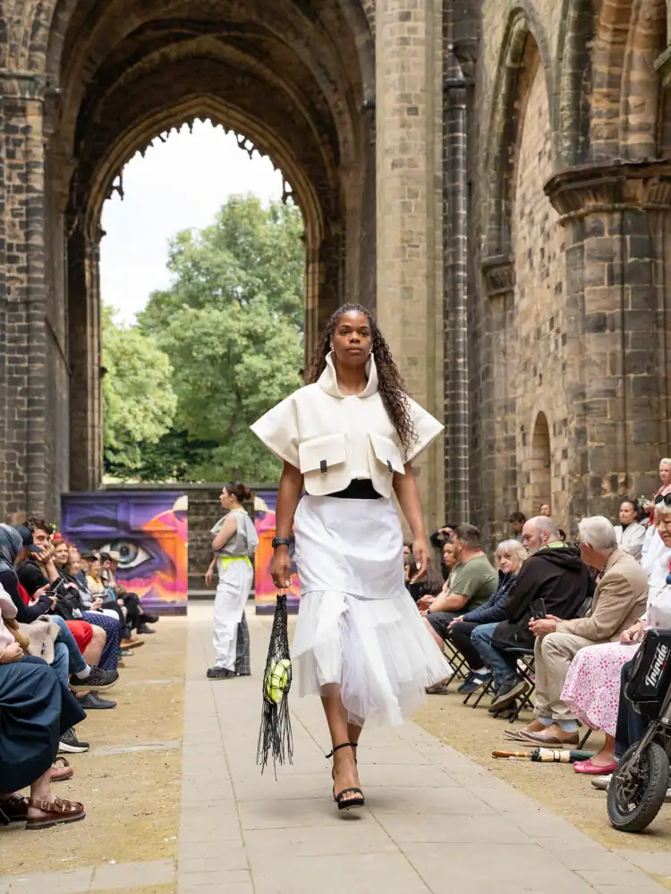 A model walks the catwalk in a cream structured cropped jacket with patch pockets, a white pleated tulle-hemmed skirt, and strappy heels, carrying a black woven bag.
