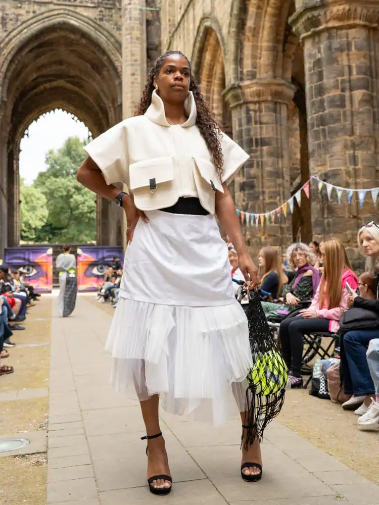 A model stands in a cream structured cropped jacket with patch pockets, a white pleated tulle-hemmed skirt, and strappy heels, carrying a black woven bag, hand on hip.
