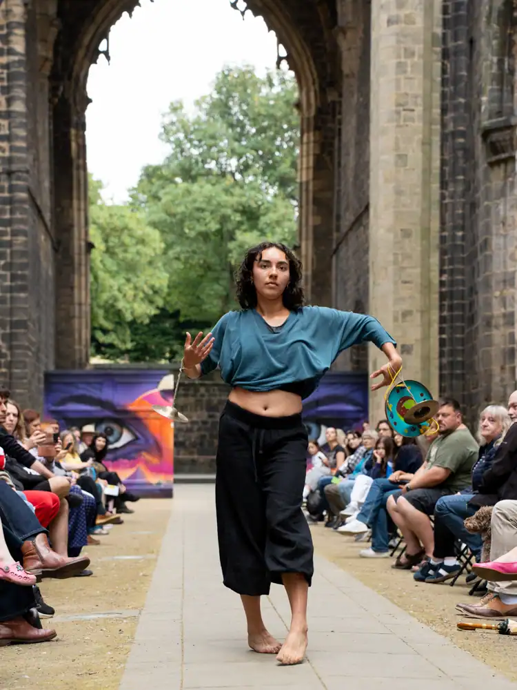A barefoot performer in a teal top and black wide-leg trousers walks with arms extended, small cymbals hanging from each hand.
