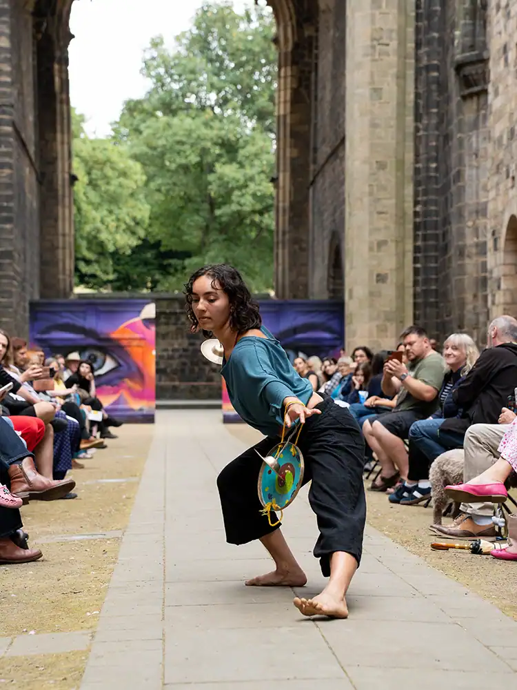 A barefoot performer in a teal top and black wide-leg trousers crouches low, reaching forward with small cymbals in hand.