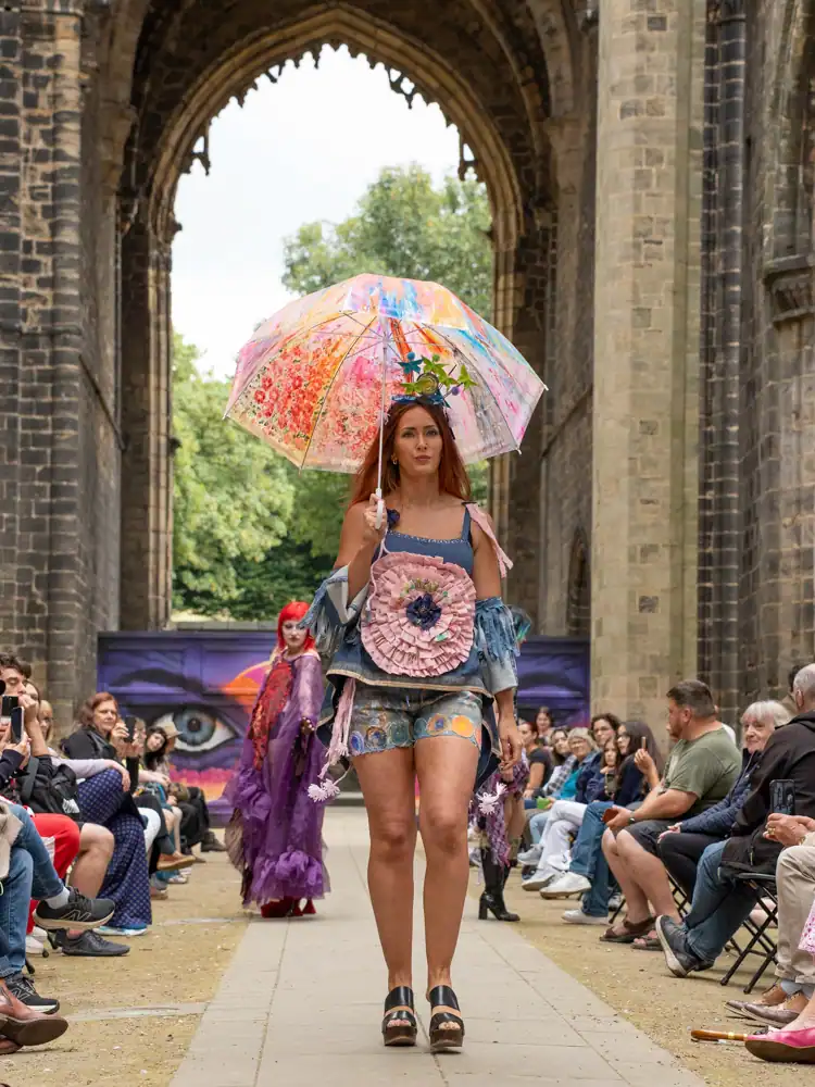 A model walks the catwalk holding an open umbrella painted with a floral design, wearing a denim outfit with a large pink ruffled flower appliqué and a floral headpiece.