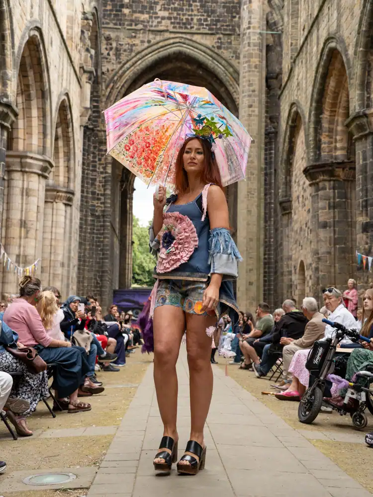 A model stands holding an open umbrella painted with a floral design, wearing a denim playsuit with a large pink ruffled flower appliqué, a floral headpiece, and platform sandals.