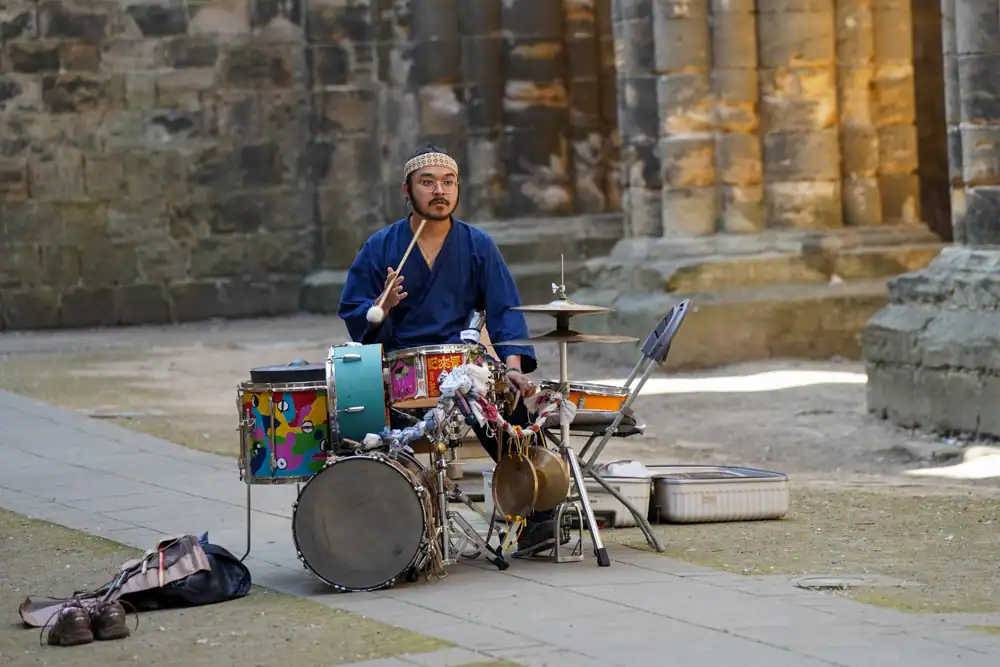 A musician in a blue robe and headscarf plays a decorated drum kit with cymbals, drumstick raised.