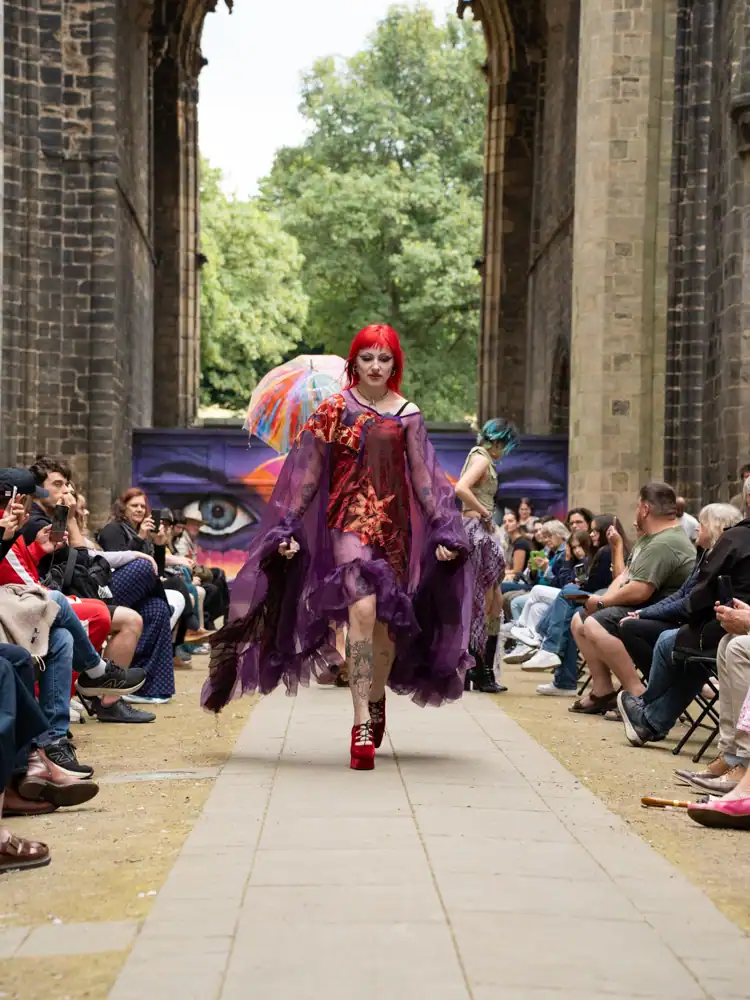A model walks the catwalk in a sheer purple ruffled cape-style dress with a red textured appliqué on the front, with red hair and platform heels.