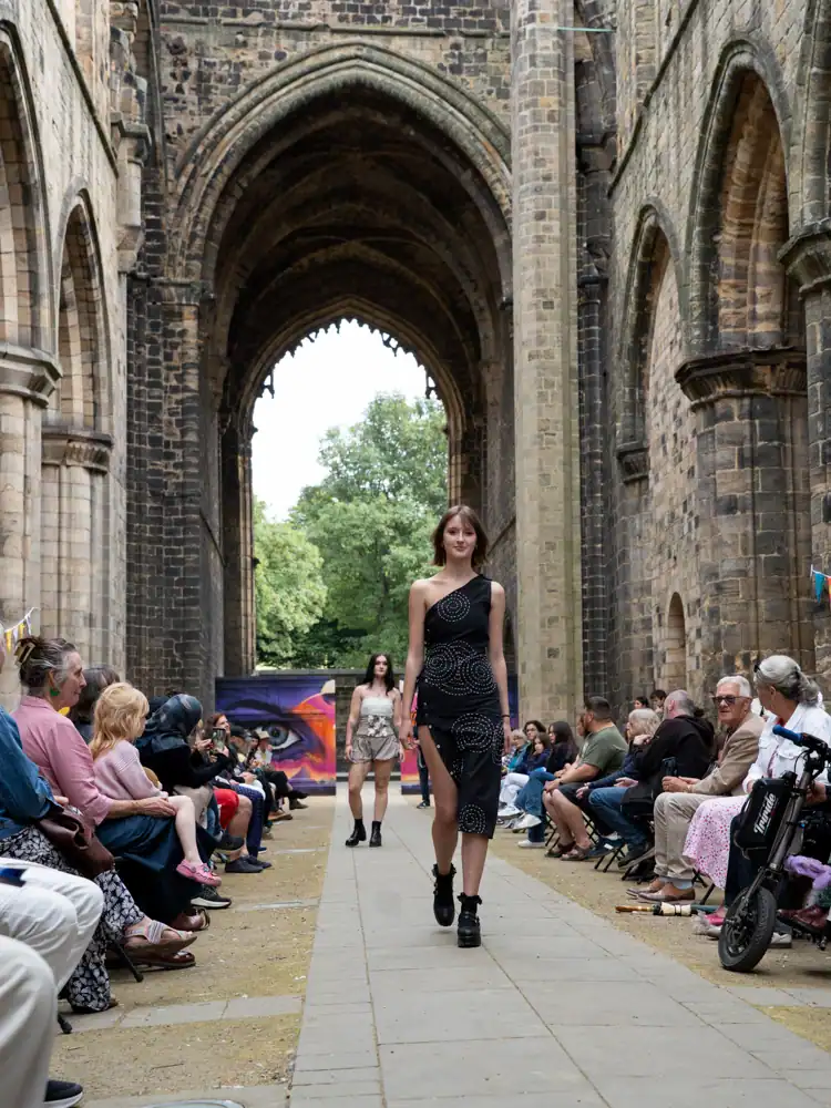 A model walks the catwalk in a black one-shoulder dress with circular beaded embroidery and a thigh-high slit, paired with black boots.