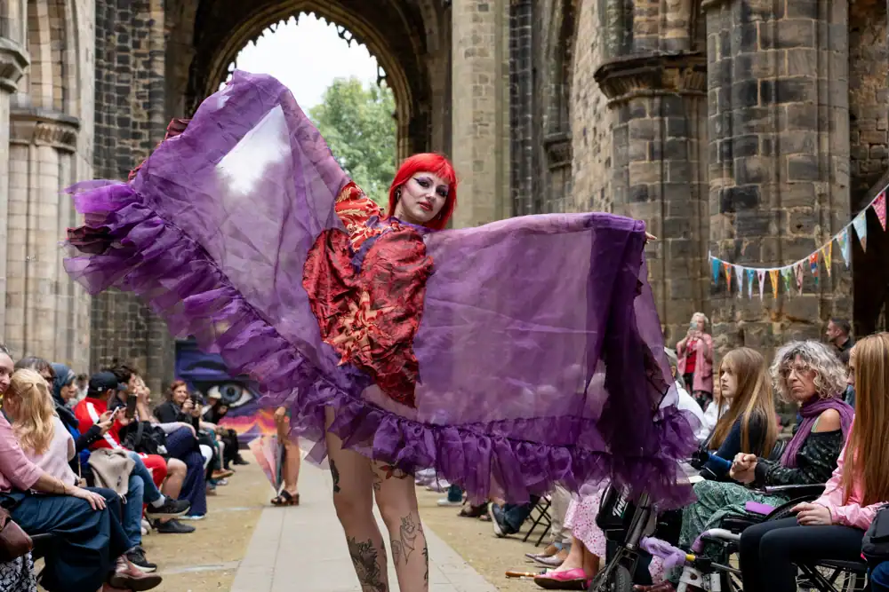 A model poses with arms spread, wearing a sheer purple ruffled cape-style dress with a red textured appliqué on the front, red hair visible, tattooed legs bare.