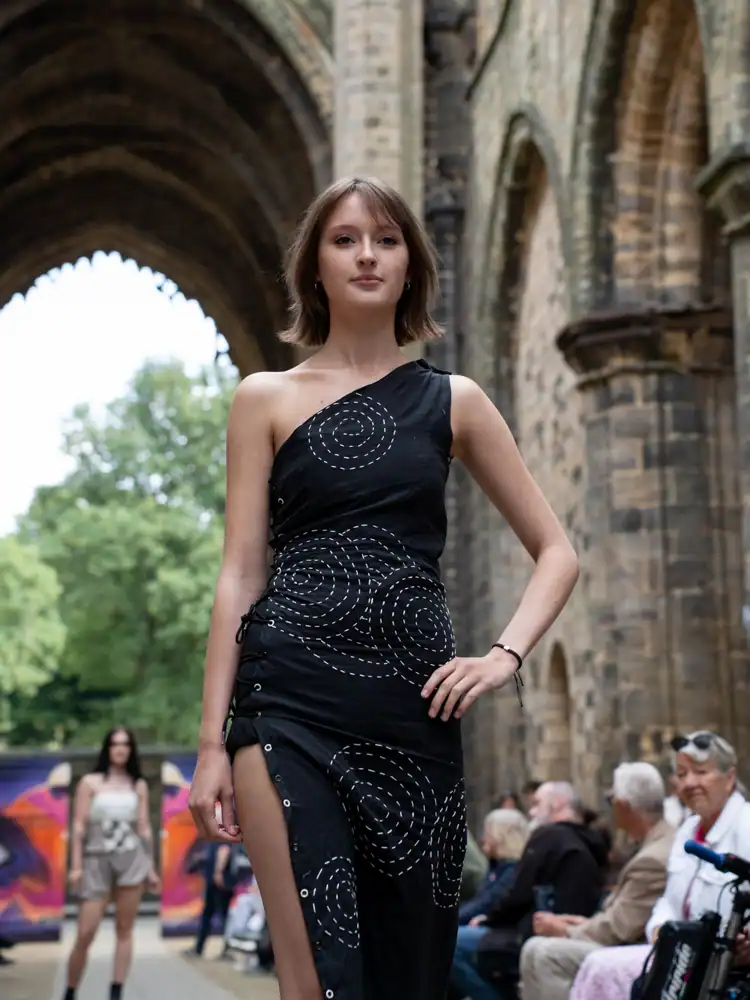 A model stands wearing a black one-shoulder dress with white spiral stitching and side lacing.