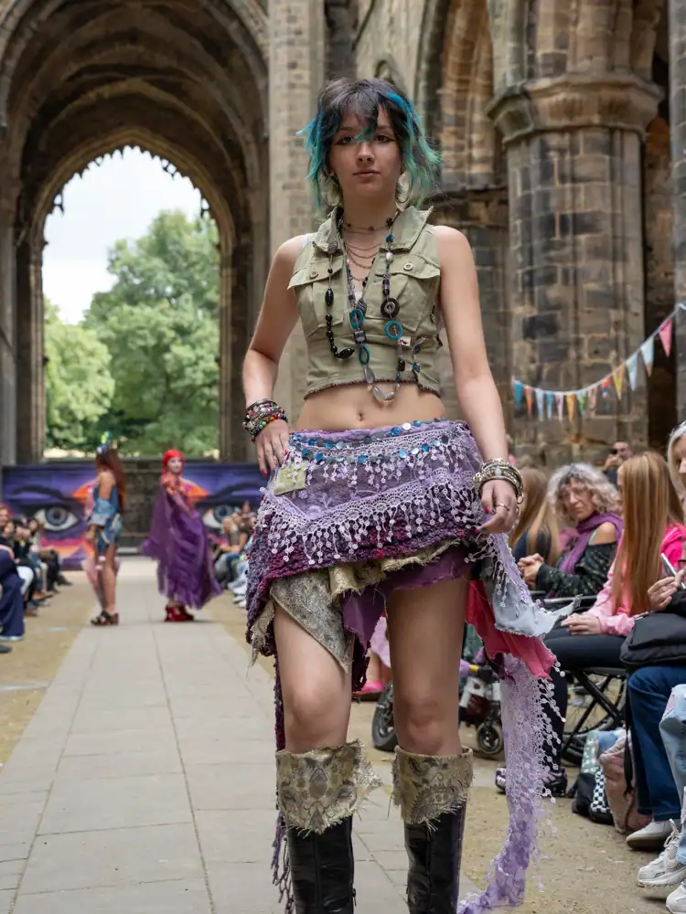 A model stands wearing a khaki cropped top with a purple lace and sequin skirt and knee-high patterned boots.