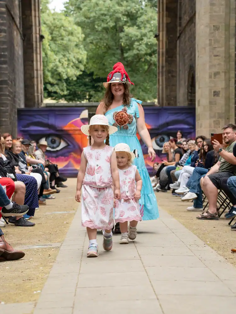 A woman and her two children modelling on the runway: the woman in a blue tiered dress with an octopus hat and crab applique, and her two children in matching pink shell-print co-ords and straw hats.