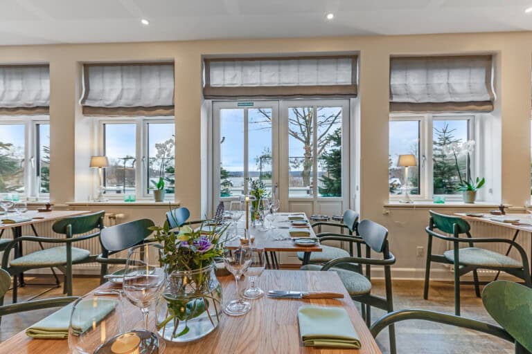 Højtbelyst restaurant med udsigt over vandet og hyggelige indretningselementer.