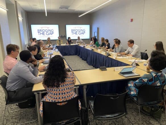 Group of individuals sitting at a square table with two TV's projecting "Bridging the Gap" on the screen.