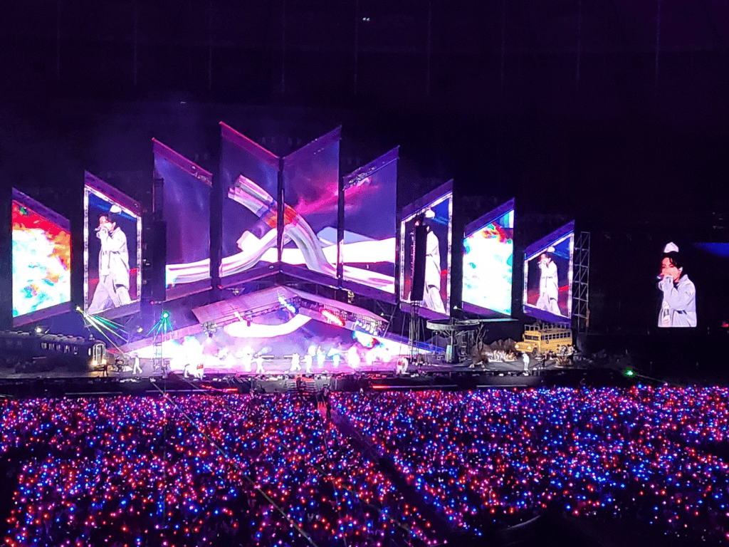 A K-Pop group performing on stage with bright lights and fans cheering.