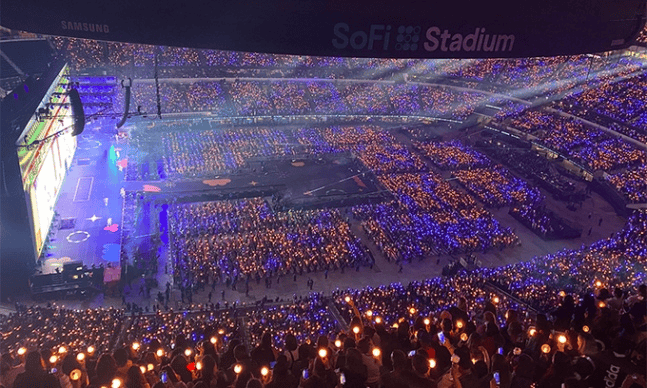 A large K-Pop concert stage with lights, screens, and thousands of cheering fans.