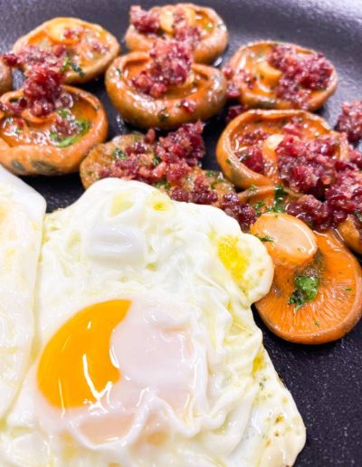 Sartén presentando dos huevos de corral fritos con puntilla y yema líquida, acompañados de un salteado de níscalos frescos y jamón en KRAM.