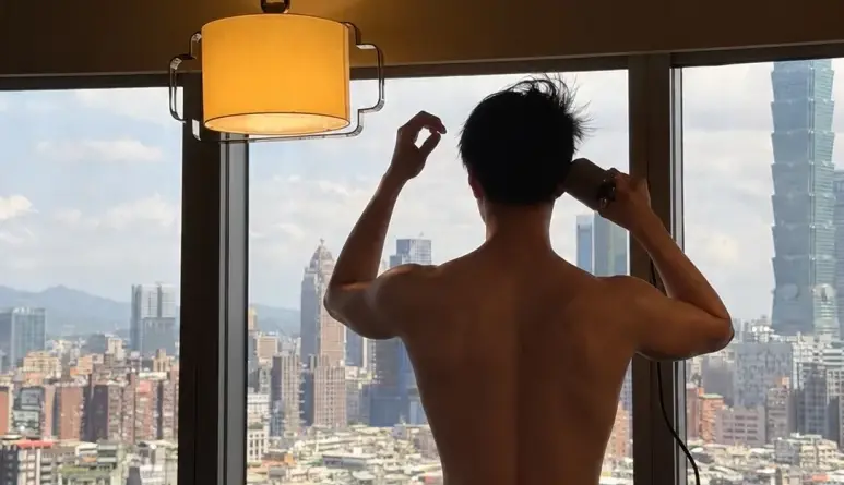 Park Bo-gum standing with his back to the camera beside a window overlooking the city