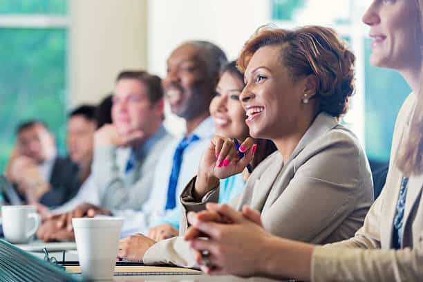 Mid adult African American businesswoman is smiling while attending a job seminar or job training business conference. She s taking notes while listening to a speaker. She is dressed in smart business casual clothing and is sitting next to diverse group of professional coworkers.