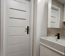 : Białe drzwi łazienkowe z czarną klamką oraz biała szafka podumywalkowa z czarną baterią.