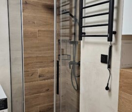 Kabina prysznicowa walk-in z czarnymi profilami, czarnym grzejnikiem i płytkami imitującymi drewno.