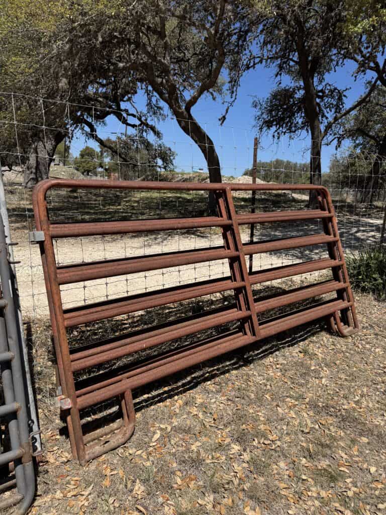 Rusty metal farm gate with multiple horizontal bars, outdoors on dirt ground.