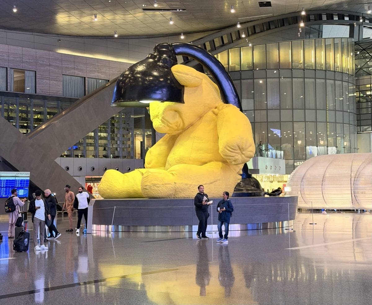 Giant plush toy of a yellow character with a black hat at an airport.