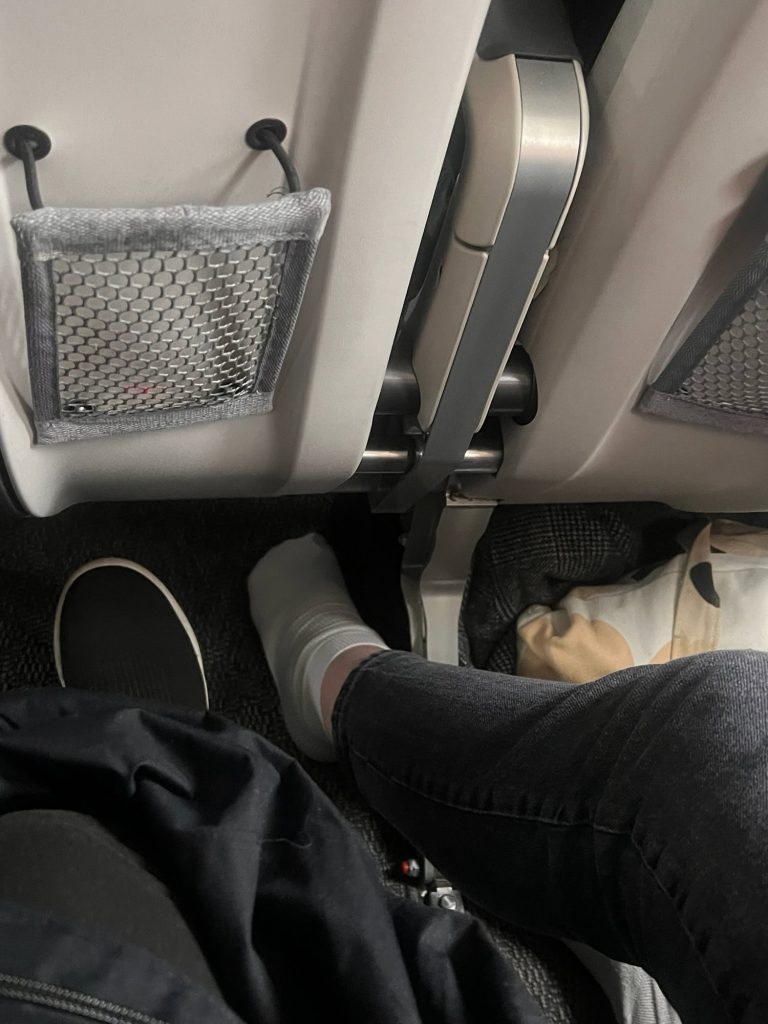 Airplane passenger seat view showing legs, seat back pockets, and personal belongings.