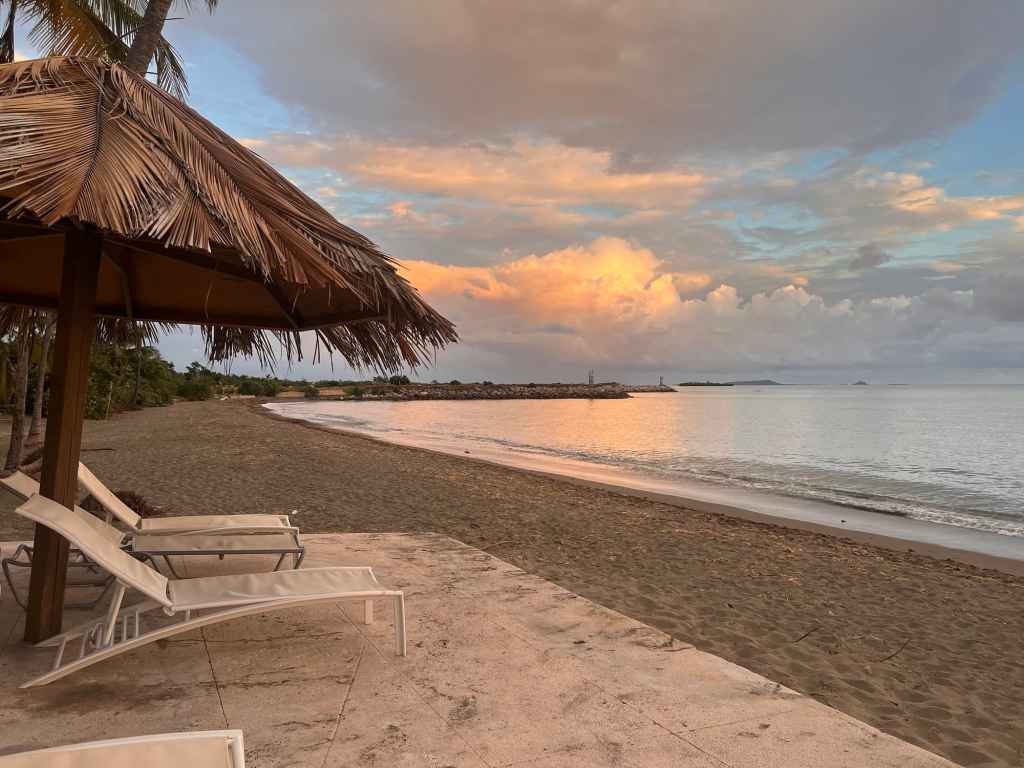 Beach sunset with lounge chairs and thatched umbrella on a sandy shore.