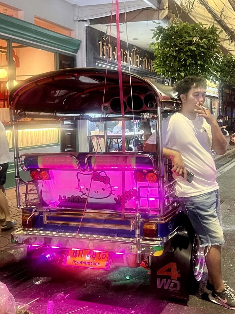 Pink Hello Kitty tuk-tuk with neon lights and a young man standing nearby in a city street.