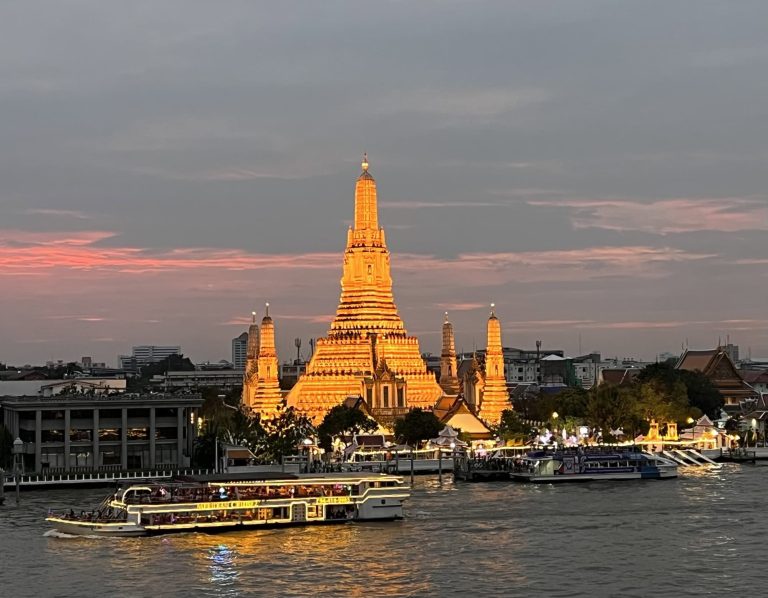 Bangkok temple illuminated at sunset with boats on the Chao Phraya River.