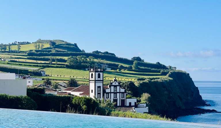 Coastal landscape featuring a white church with black accents, lush green hills, and the ocean in th.