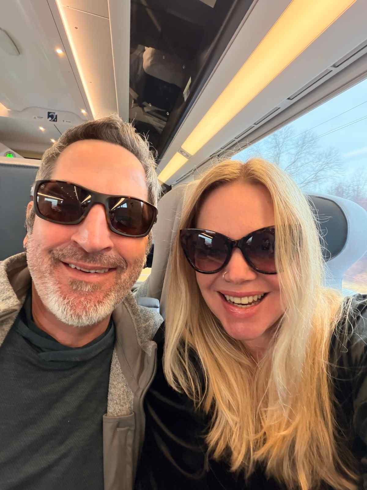 Travel couple on train smiling, wearing sunglasses, during their journey.