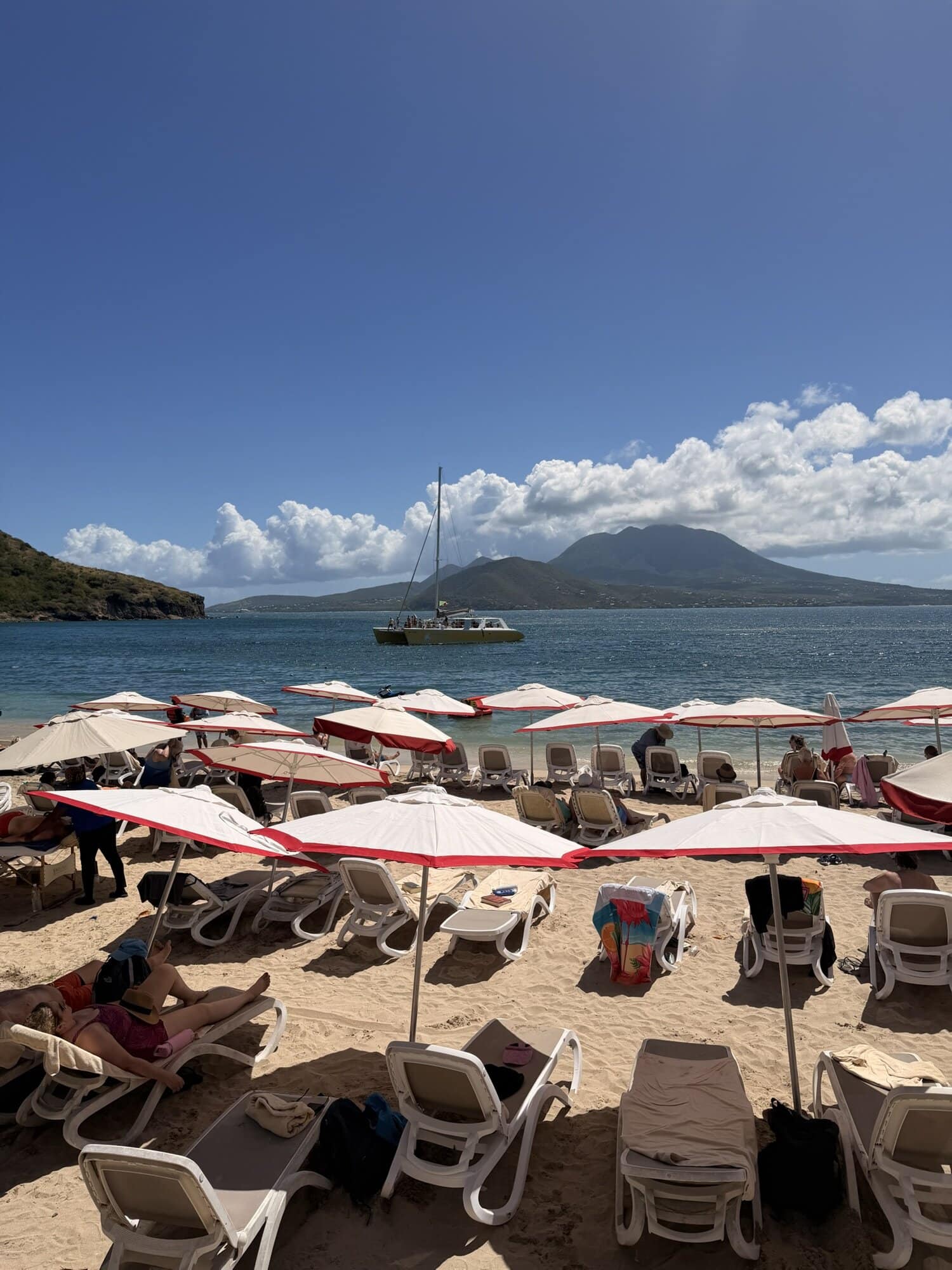 Beach with umbrellas and lounge chairs, sailboat on the water, mountain in the background.