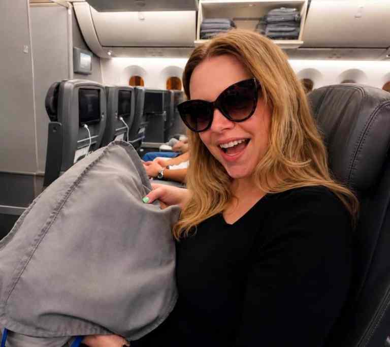 Woman with sunglasses smiling on airplane, holding a pillow, enjoying travel experience.