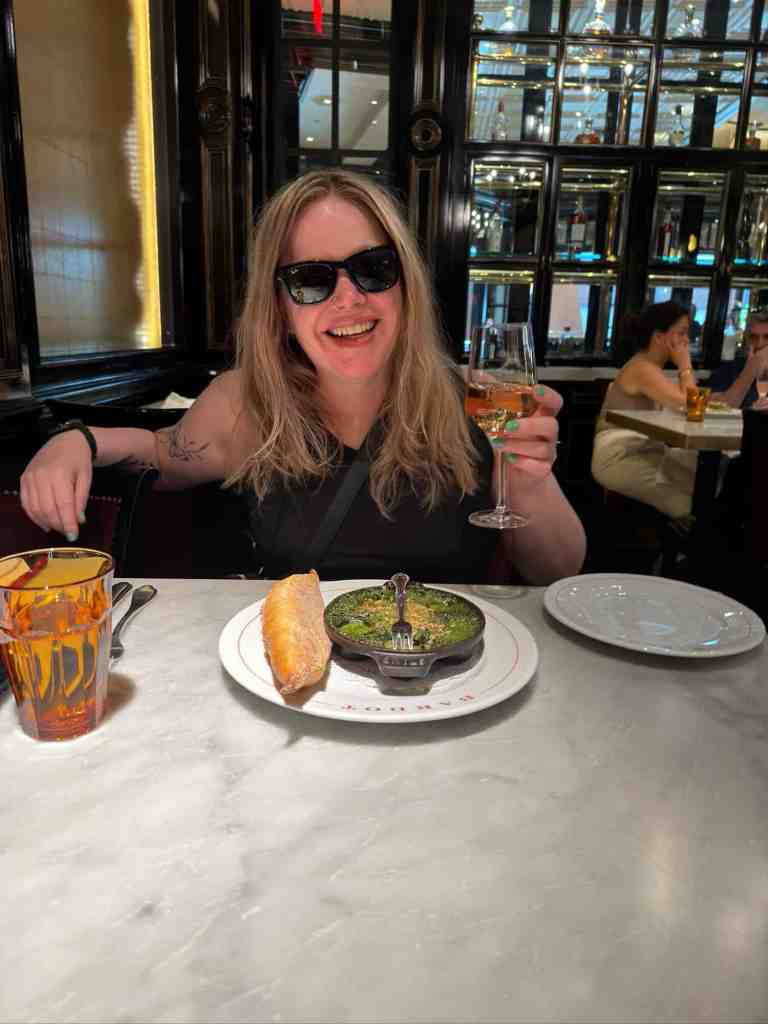 Woman with sunglasses smiling, holding a glass of wine, seated at a table with French cuisine.