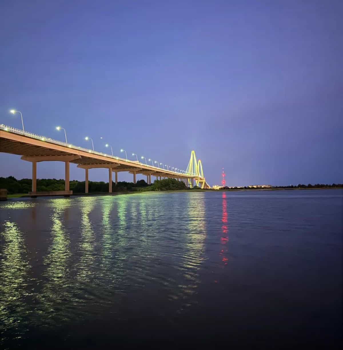 Night view of a lit-up bridge over water with city lights in the distance.
