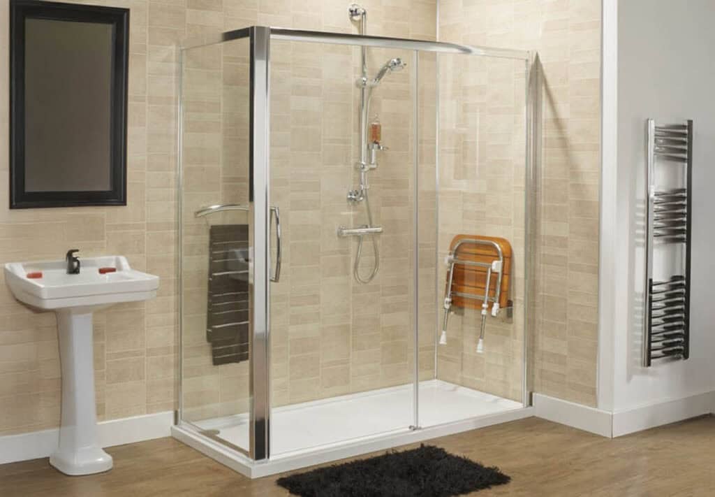 Premier accessible bathroom with glass shower enclosure, adjustable wooden seat, pedestal sink, and chrome towel warmer on beige tiled walls.