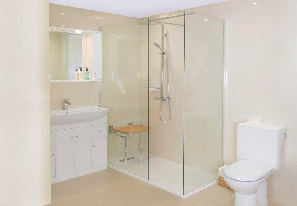 Accessible bathroom with glass-enclosed shower, wooden fold down bench, wall-mounted shower head, and white vanity with mirror.