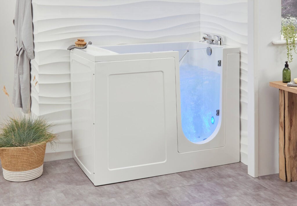 Walk in shower bath combination Milano (Georgia). Filled with water, illuminated baby blue by light and bubbles from jets. White walls carved into waves behind walk in bath.
