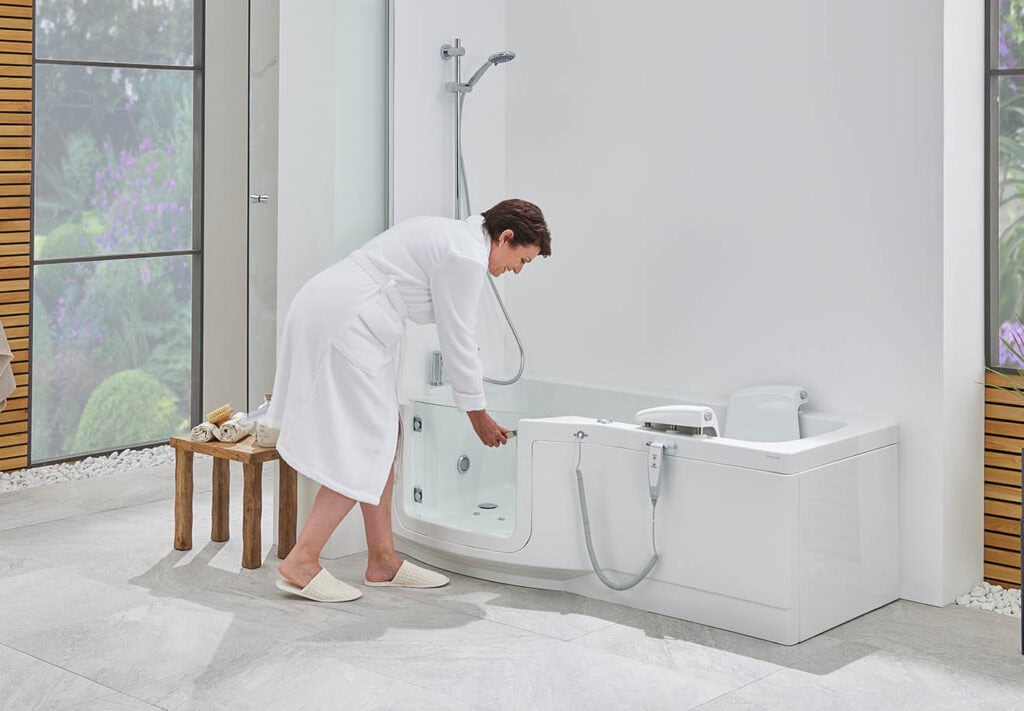 Roma Stretto (indiana) walk-in shower bath. elderly lady wearing white robe, learning in to open the glass door. bright white open plan bathroom with large wall size window.