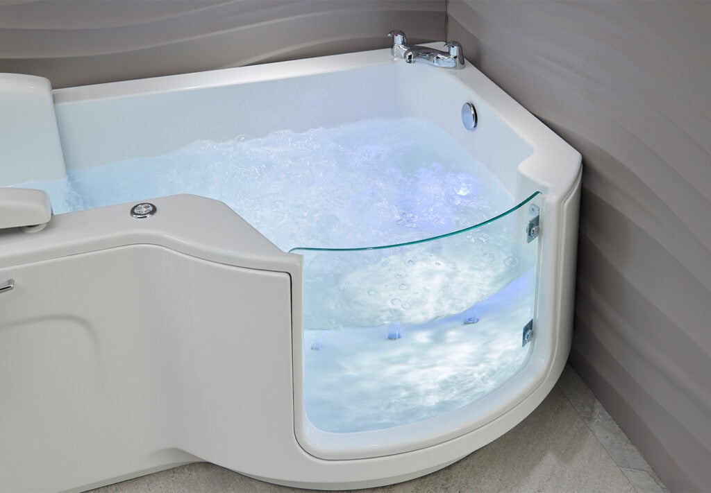 Walk-in bath Roma (Louisiana - access baths) zoomed in half view bath glass door, filled with water and bright blue light illuminating inside of tub. In neutral coloured theme bathroom.