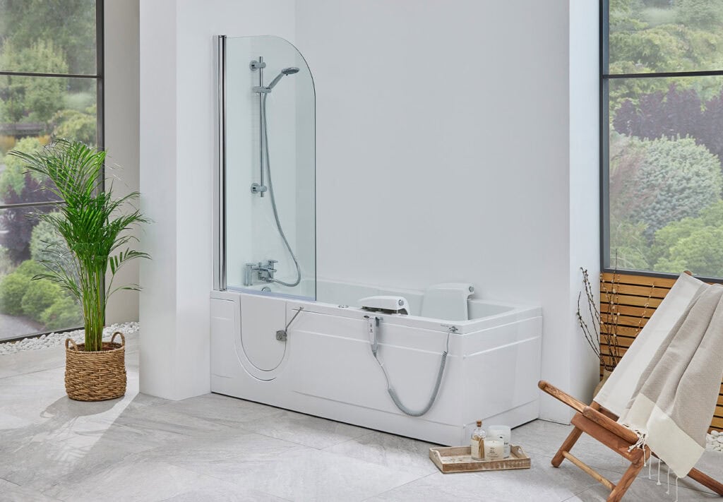 Livorno Supreme (Montana) walk in shower bath. Bright white mobility bathroom. Wood sun lounger chair with towel draped over next to bath and tall plant in weaved wooden pot on other side of bath.