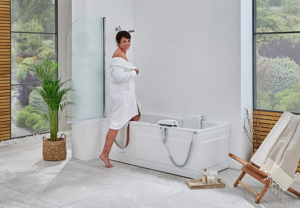 Elderly women stepping into Livorno Supreme (Montana) walk in shower bath. Bright white mobility bathroom. Wood sun lounger chair with towel draped over next to bath and tall plant in weaved wooden pot on other side of bath.