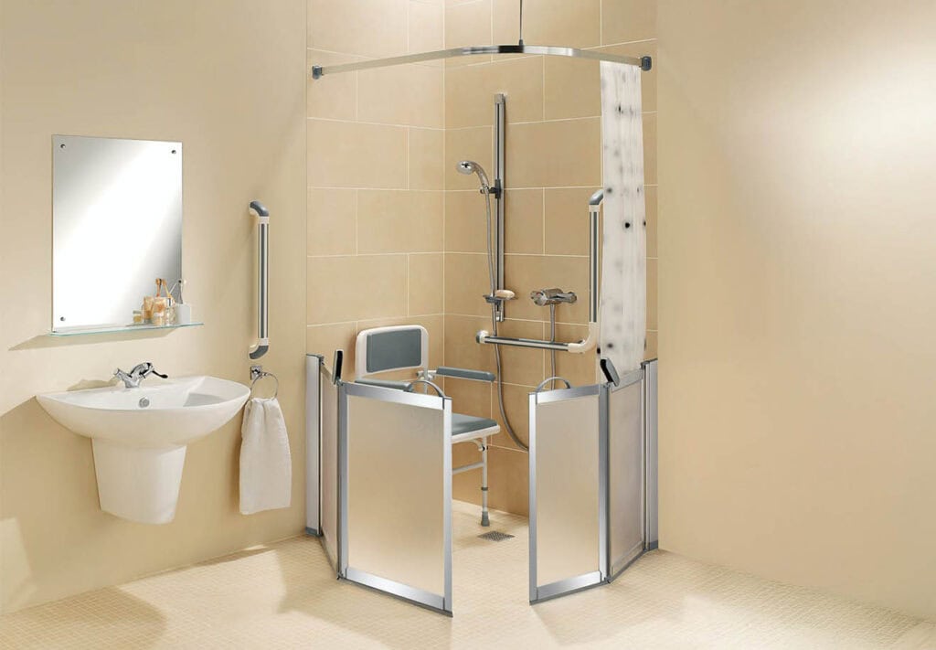 Accessible bathroom with half-height shower doors, foldable chair, grab bars, handheld shower head, and adjacent sink with mirror.