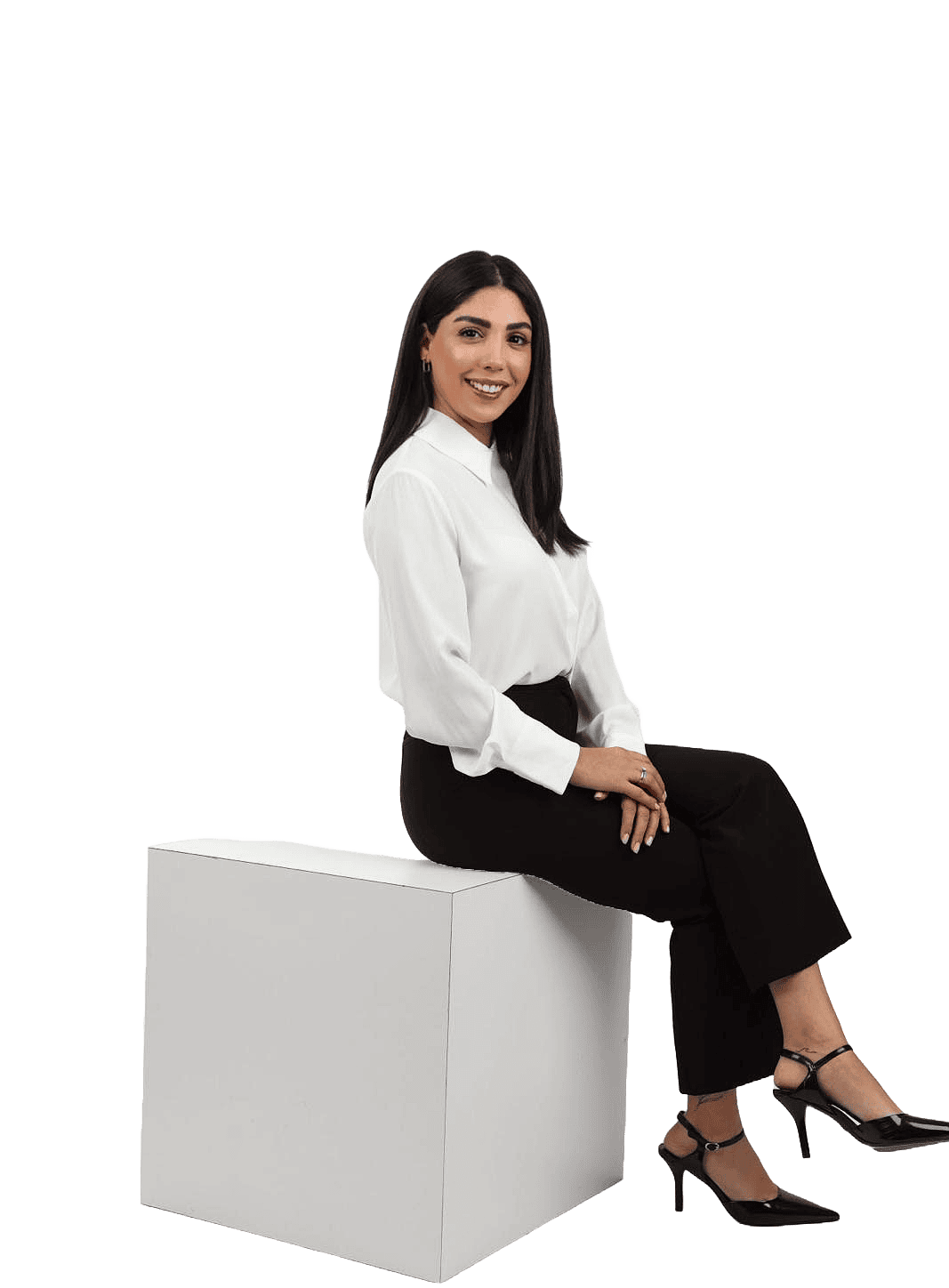 אישה מחייכת, לבושה בחולצה לבנה ומכנסיים שחורים, יושבת על קובייה לבנה.