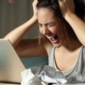 A young woman, beset by laptop and chaos&mdash;hair-grabbing, mid-scream, surrounded by crumpled papers and a turquoise pencil case.