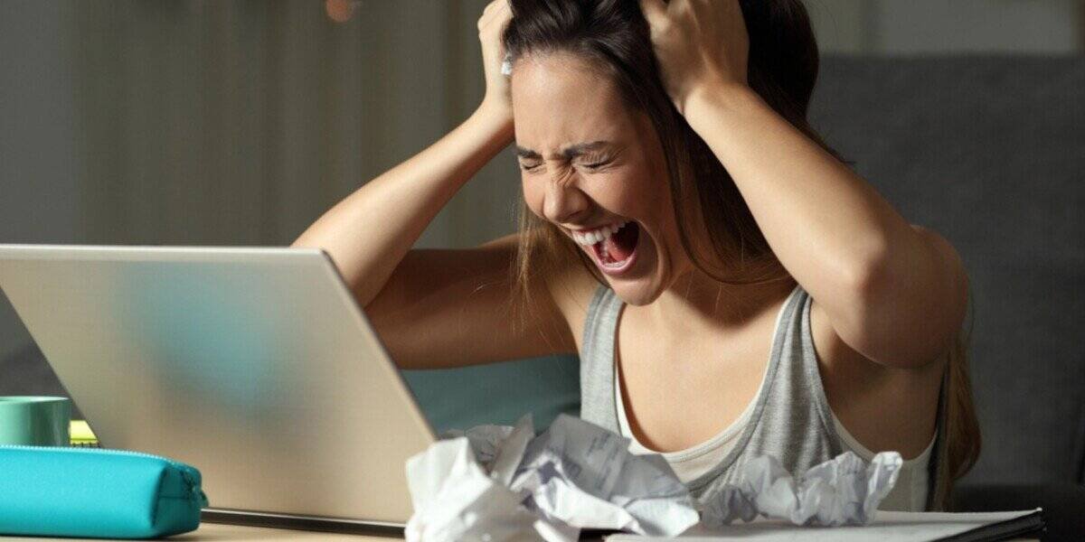A young woman, beset by laptop and chaos—hair-grabbing, mid-scream, surrounded by crumpled papers and a turquoise pencil case.