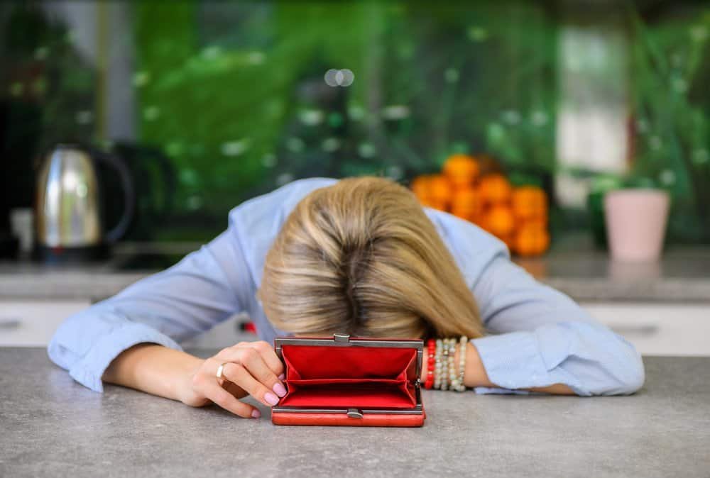 A blonde woman slumped over the kitchen worktop, clutching an empty red purse as if hoping it would magically refill, while oranges, a kettle, and the pink cup watched in silent solidarity.