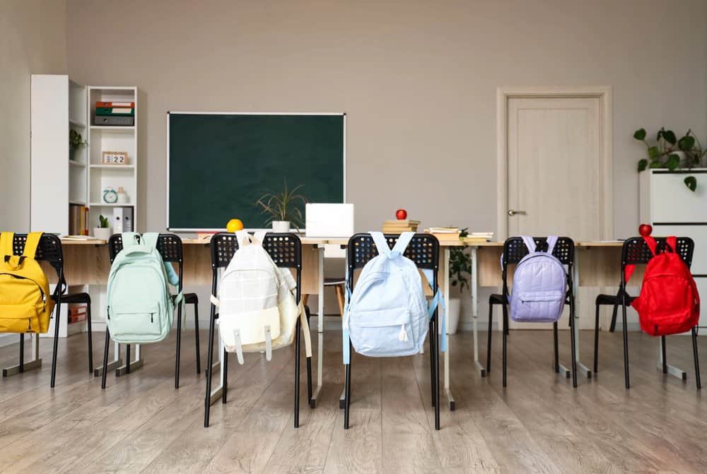 A classroom, clearly between invasions of small humans, sits in patient anticipation. Black chairs—so regimented, so empty—each parade a rucksack as flamboyant as a wizard’s hat at festival time. Desks groan gently beneath the weighty expectation of books (for knowledge), apples (for hopeful bribery), and supplies (for unspeakable arts and crafts). At the rear stands a green blackboard, wider than an Unseen University dining table; plants disguise themselves as decor but are unmistakably plotting escape; shelves guard their mysterious educational treasures. The vast expanse of pale wooden floor gleams under light that suggests magic—or at least very clean windows.