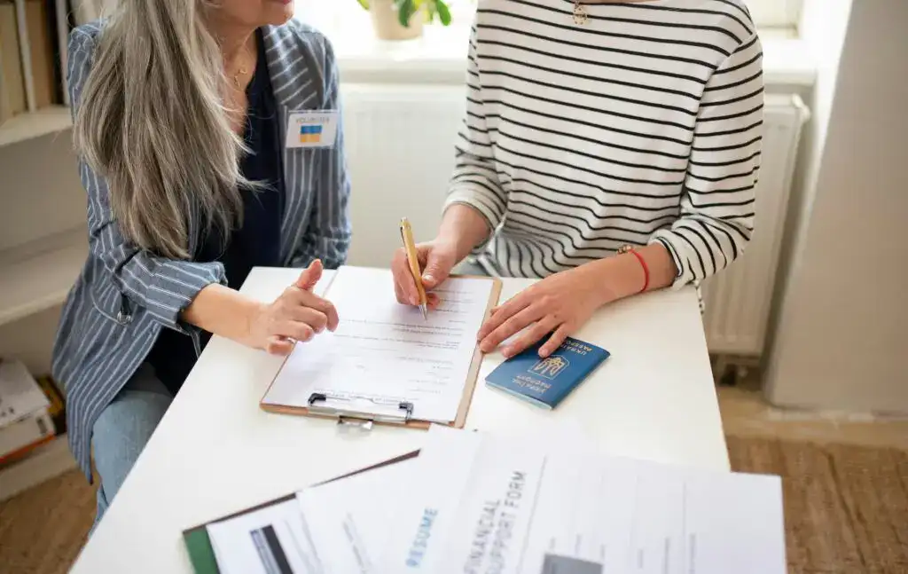 Two people sitting at a table reviewing and signing official documents, with a passport and paperwork visible.