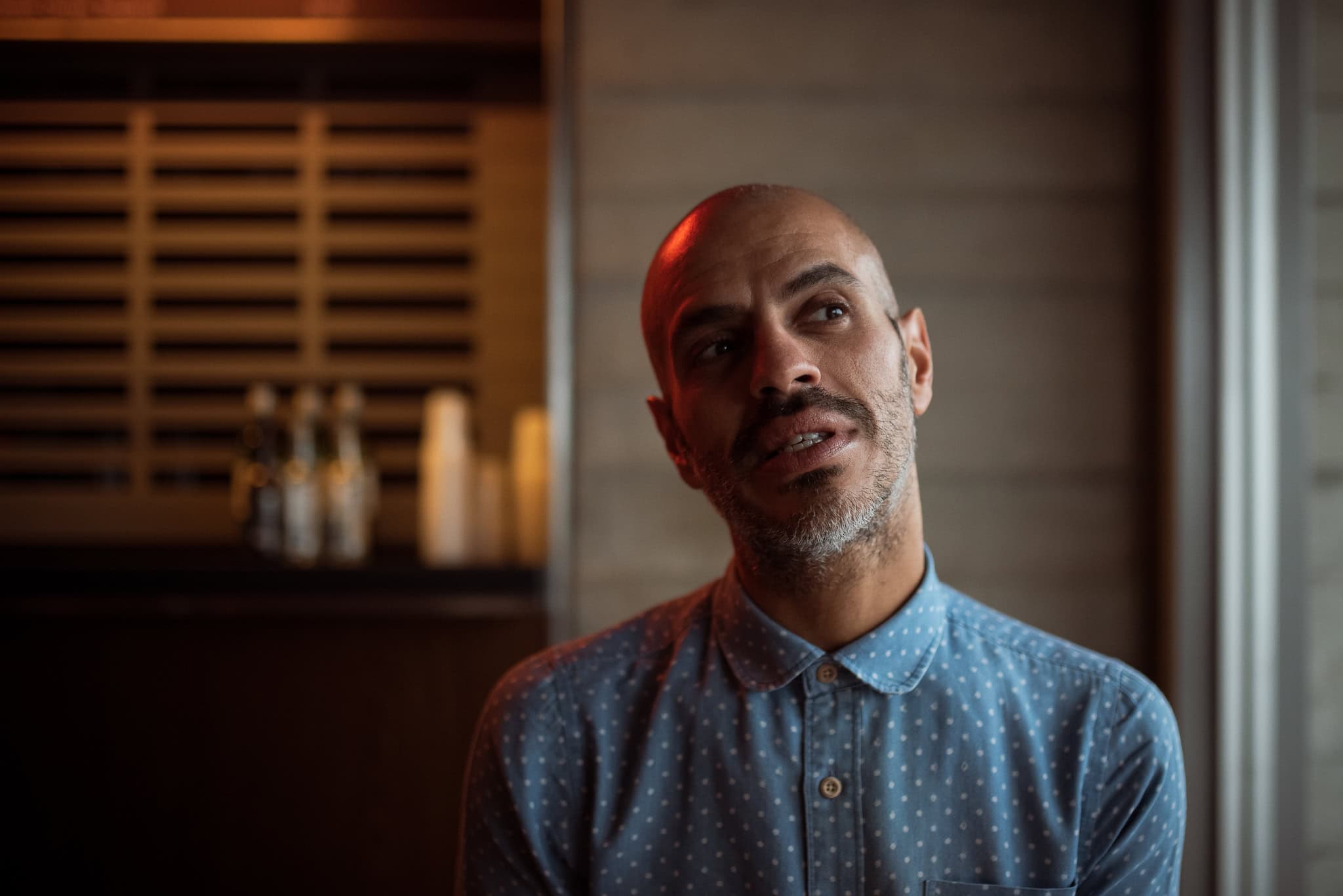 Cahit Ali, a man with a shaved head and beard, wearing a blue patterned shirt, looks thoughtfully upwards in soft lighting.
