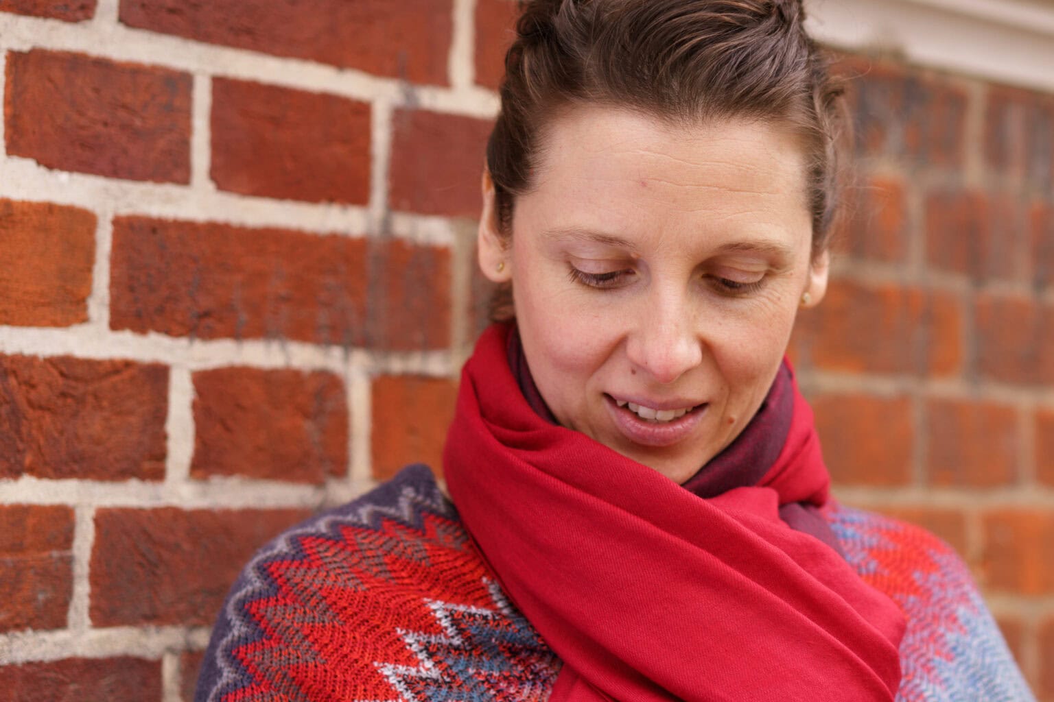 Moni Soos, a coach, looks down with a warm smile, wearing a red scarf and patterned jumper against a brick wall. London brand photographer.